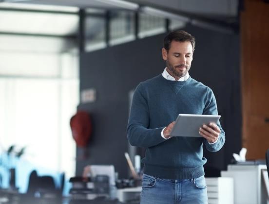Person in blue sweater and jeans using a tablet in an office with desks and chairs; face pixelated for privacy.