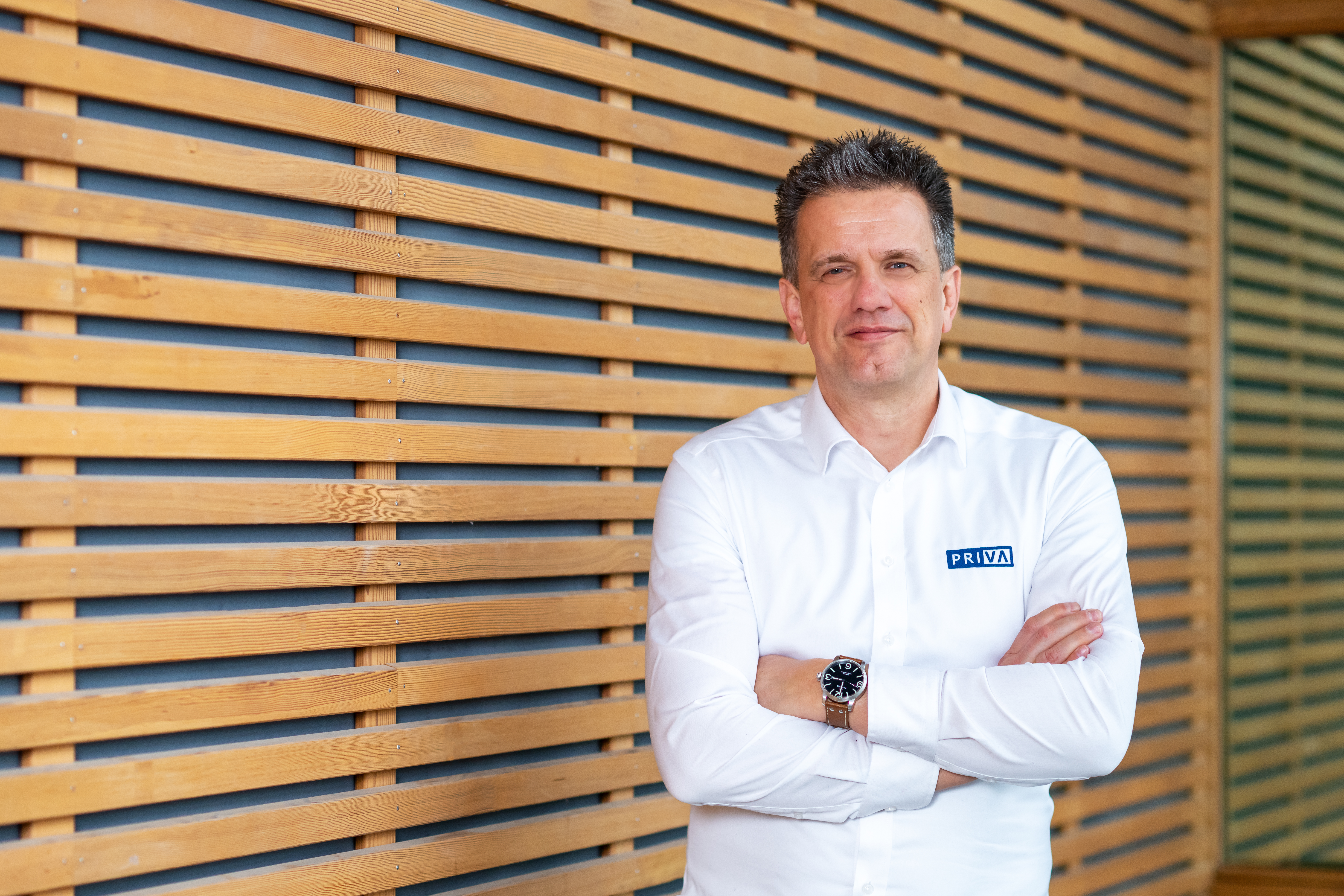 Personne portant une chemise blanche Priva, debout bras croisés devant un mur en lattes de bois.
